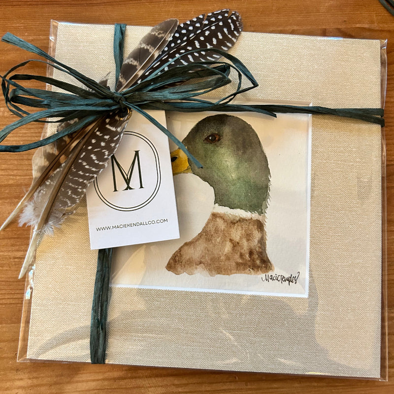 Gift wrapped in beige paper with a bird illustration, tied with green ribbon and feathers on a wooden surface.