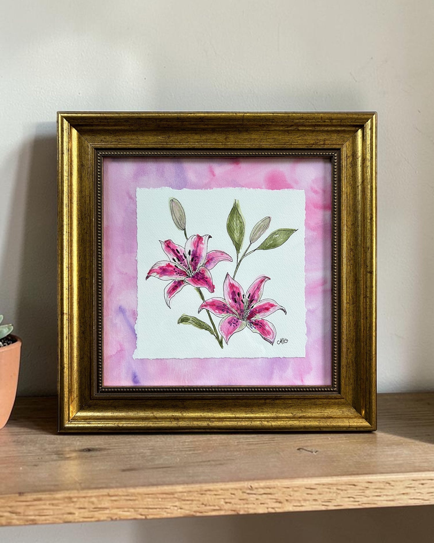 Framed artwork of watercolor and ink pink lilies on a wooden shelf with a neutral background